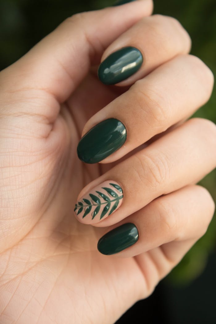 Usługi Close-up of a stylish green manicure with a leaf design, showcasing glossy fingernails and natural skin tone.