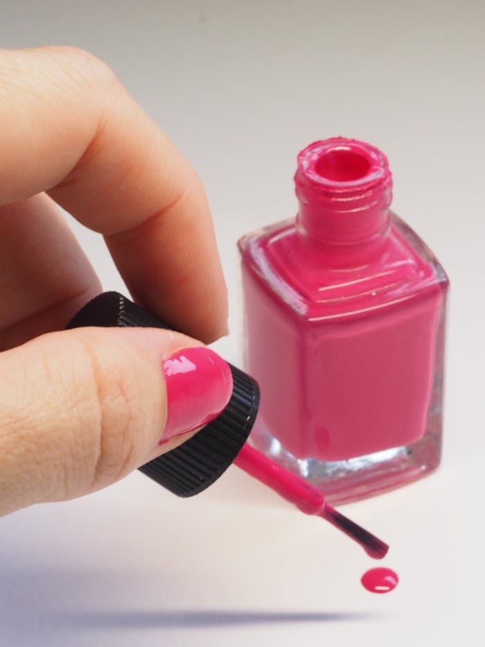 Usługi Female hand applying bright pink nail polish from open bottle, minimalistic style.